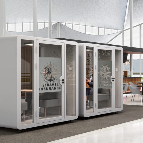 Two 4-person Quad soundproof booths side by side in an airport walkway near the boarding area, without people, displaying the logo of a travel insurance company.